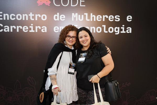 Duas mulheres sorrindo abraçadas em um evento, usando crachás, com painel