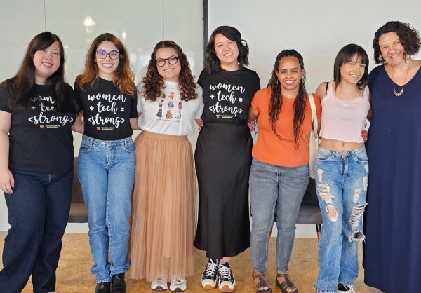 Mulheres em pé posando pra foto, todas estão abraçadas ao lado da outra