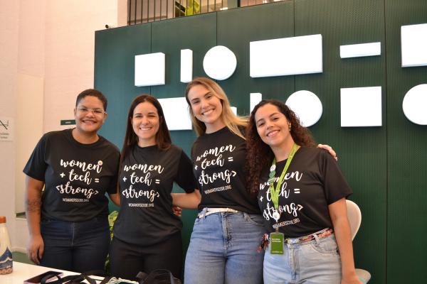 Quatro mulheres sorrindo lado a lado, usando camisetas “women + tech = strong”, em um ambiente interno de evento.