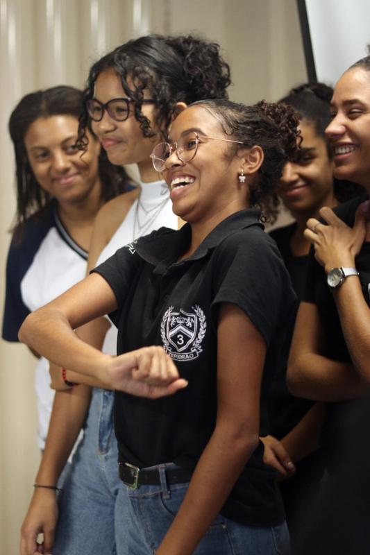 Estudantes sorridentes em grupo; ao centro, uma jovem de óculos e camisa polo preta.