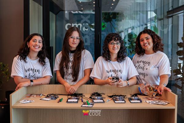 Quatro mulheres sorridentes em pé atrás de uma mesa de registro, todas usando camisetas women + tech