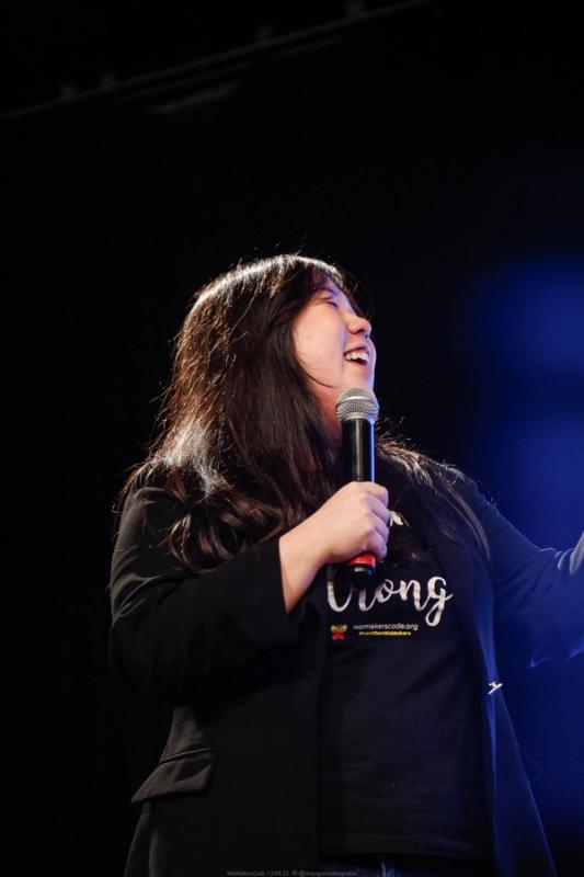 Mulher no palco segurando um microfone, sorrindo enquanto fala, com fundo escuro e iluminação de evento.