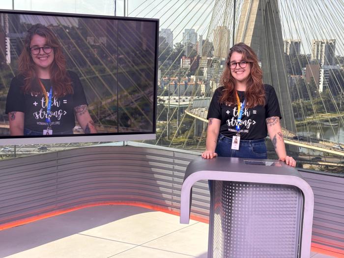 Mulher sorridente em estúdio de TV, com a Ponte Estaiada ao fundo.