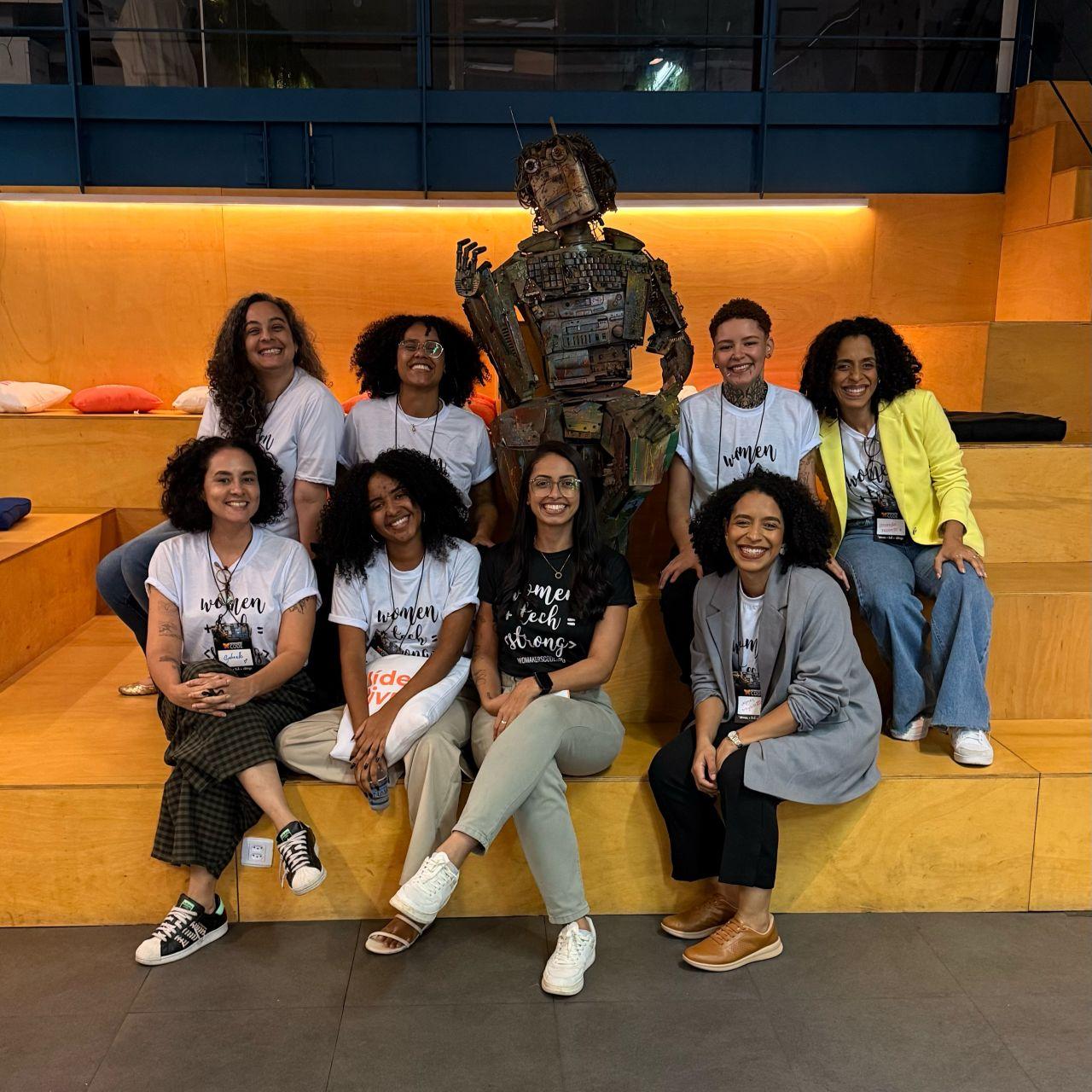 Oito mulheres sorridentes em arquibancada com escultura de robô de sucata. Elas usam camisetas "Women in Tech = Strong".