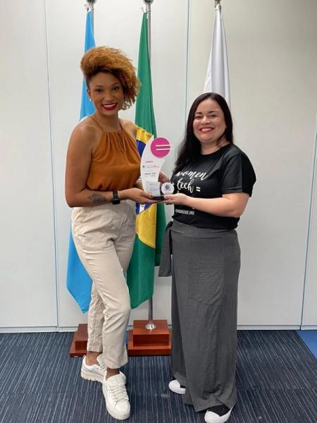 Duas mulheres sorriem e seguram um troféu juntas em frente às bandeiras da ONU e do Brasil em um escritório.