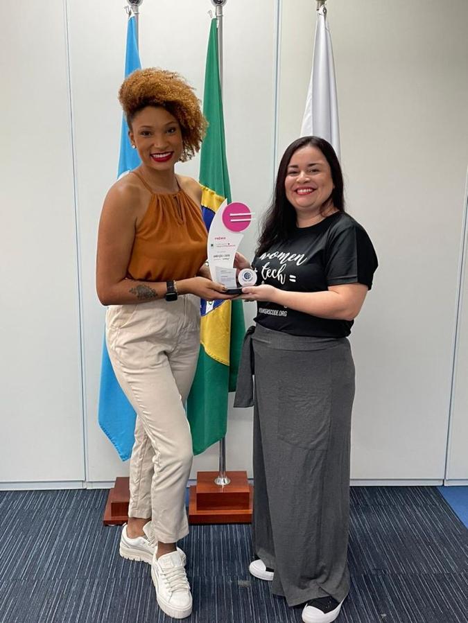 Duas mulheres sorriem e seguram um troféu juntas em frente às bandeiras da ONU e do Brasil em um escritório.