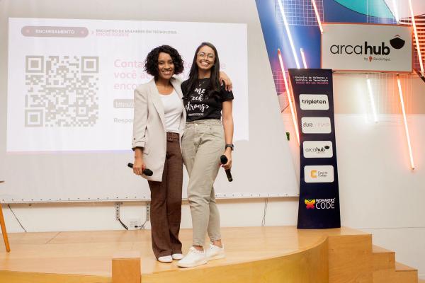 Duas mulheres sorrindo no palco de um evento, segurando microfones, com tela de apresentação e logos ao fundo.