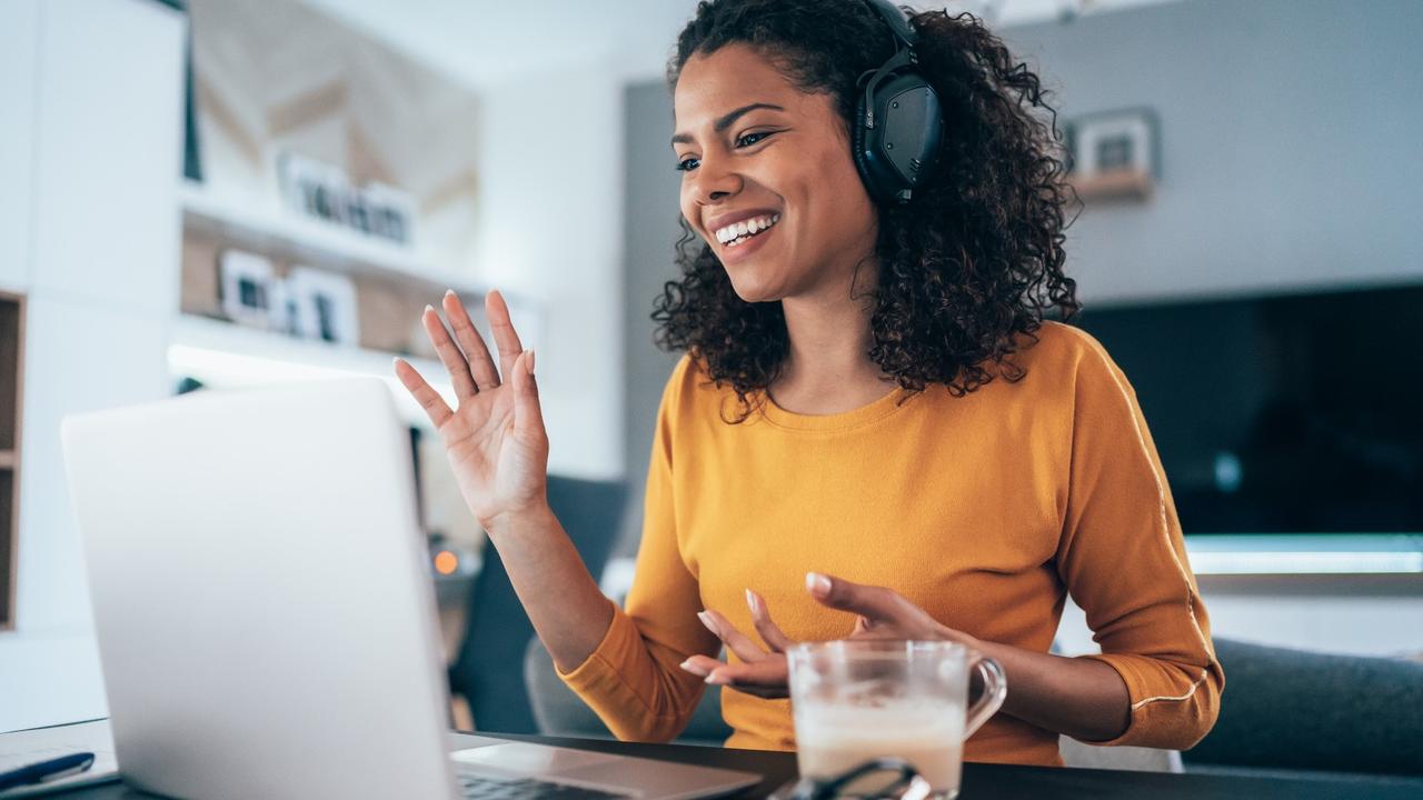A smiling woman wearing headphones waves during a video call on her laptop.