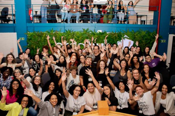 Grupo grande de mulheres reunidas em um evento, sorrindo e levantando as mãos