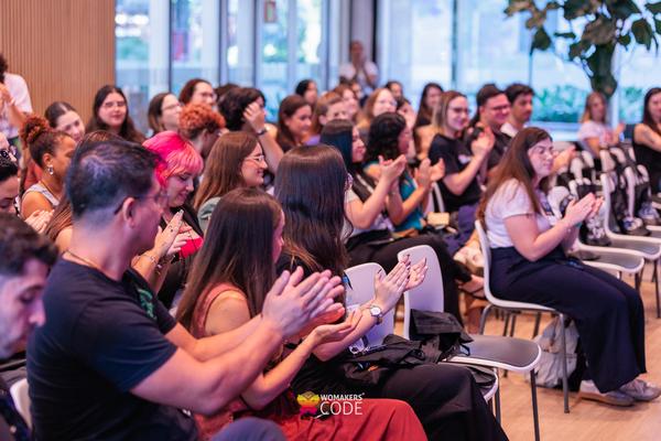 Público sentado em um evento, aplaudindo, em ambiente interno com cadeiras organizadas e clima de apresentação.