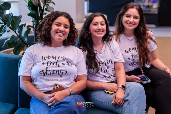 Três mulheres sentadas lado a lado, sorrindo, usando camisetas “women + tech = strong”, em um ambiente interno
