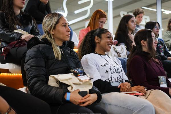 Público sentado em arquibancada durante um evento, atento à apresentação, com algumas pessoas
