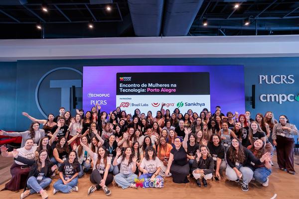 Grupo de mulheres acenando em evento de tecnologia no Tecnopuc. Ao fundo, telão: Encontro de Mulheres na Tecnologia