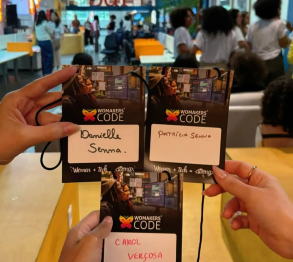Mãos seguram três crachás da WoMakersCode com os nomes. Fundo de um evento.