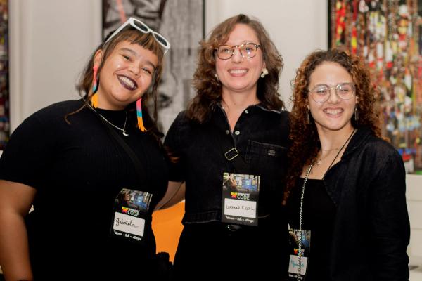 Três mulheres sorrindo, lado a lado, usando crachás da WoMakersCode em um evento, com fundo desfocado e colorido.
