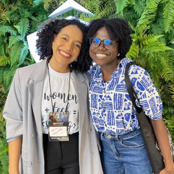 Duas mulheres negras sorridentes em um evento de tecnologia. Uma veste blazer cinza e a outra, camisa azul estampada.