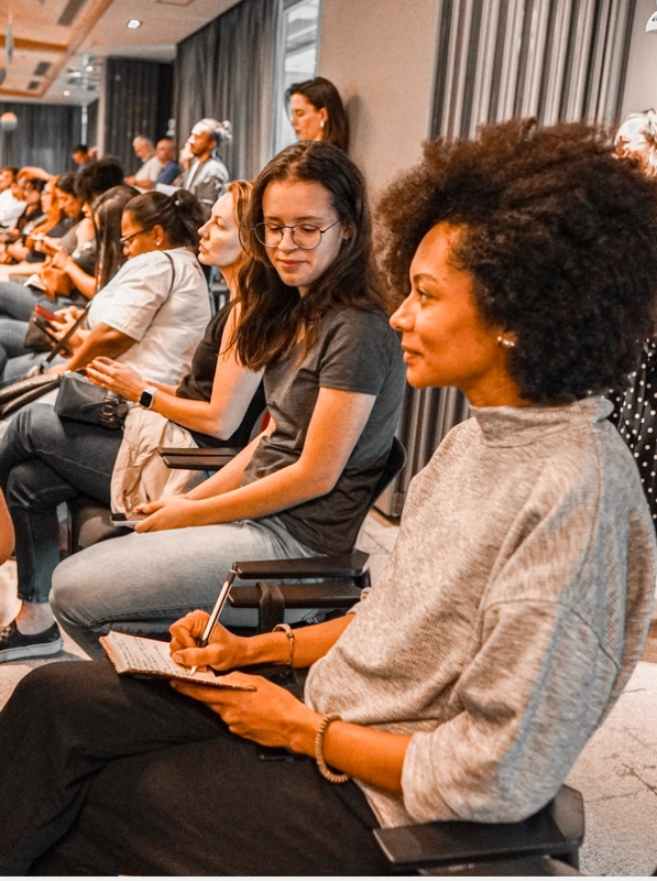 Mulher negra faz anotações em um evento. Ao seu lado, uma jovem de óculos observa atentamente.