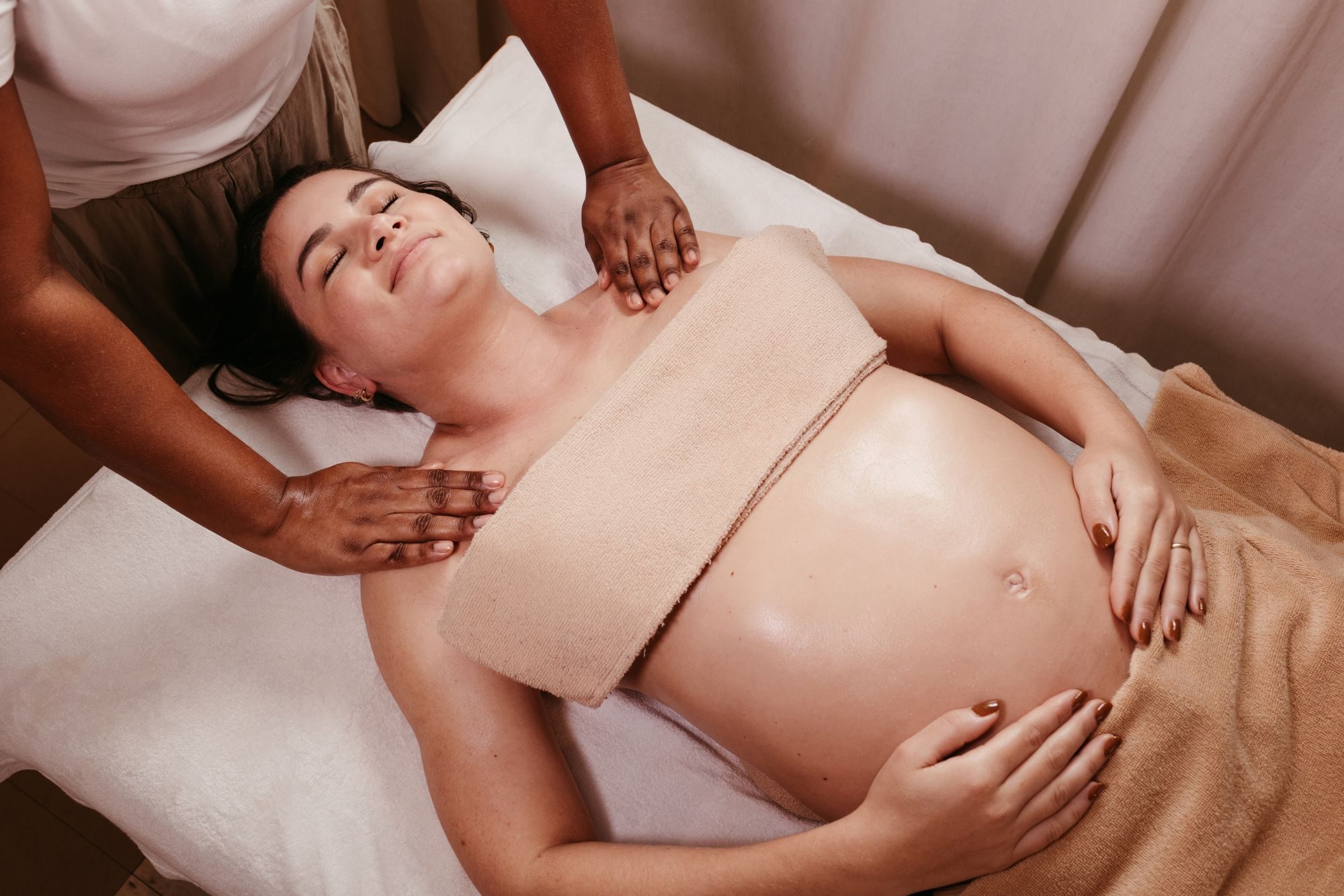 A pregnant woman rests on a massage table with her hands on her bump while a therapist massages her shoulders