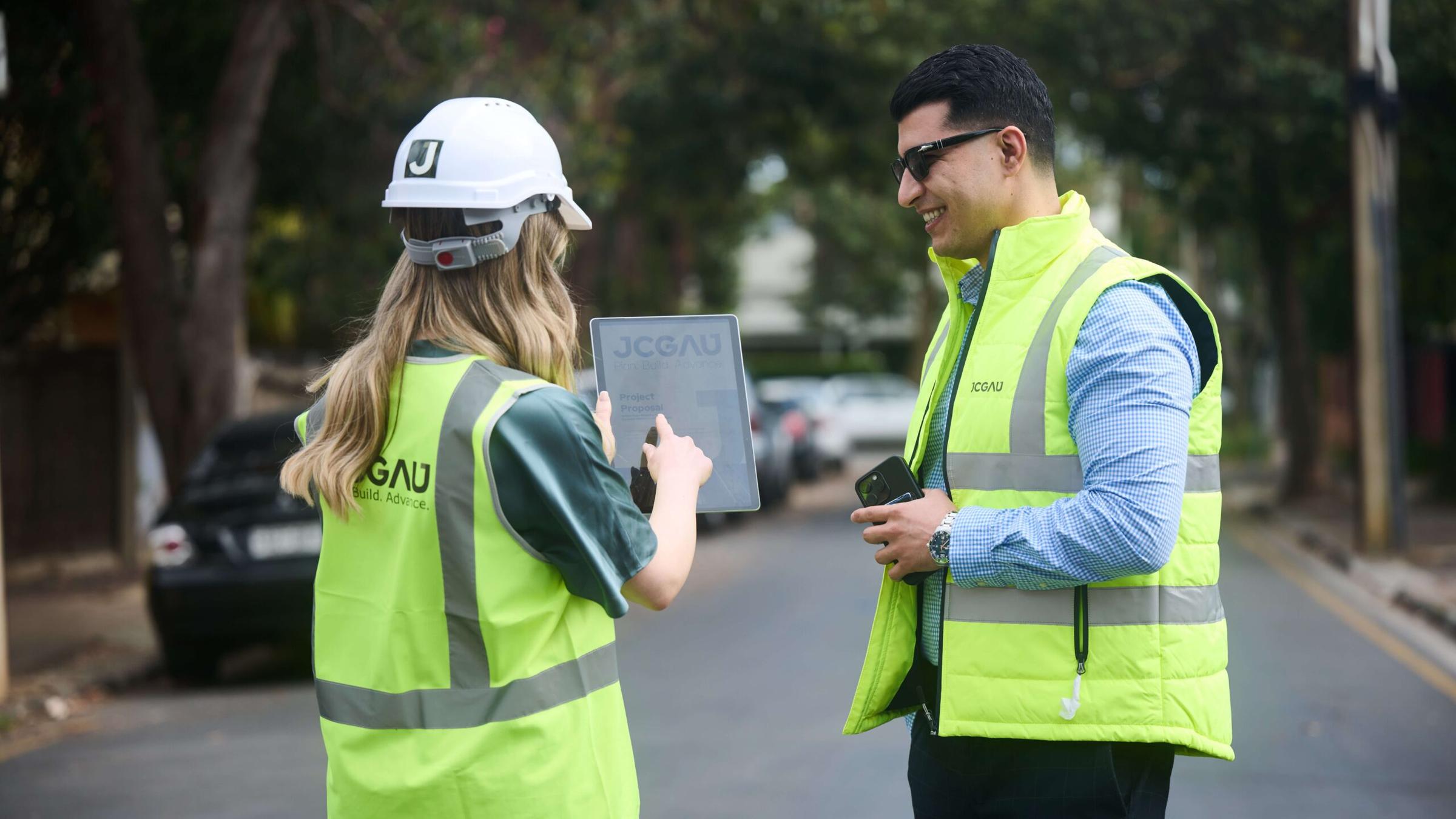 Two members of the JCGAU team on site in hi-vis vests, one in a JCGAU hard hat holding a tablet