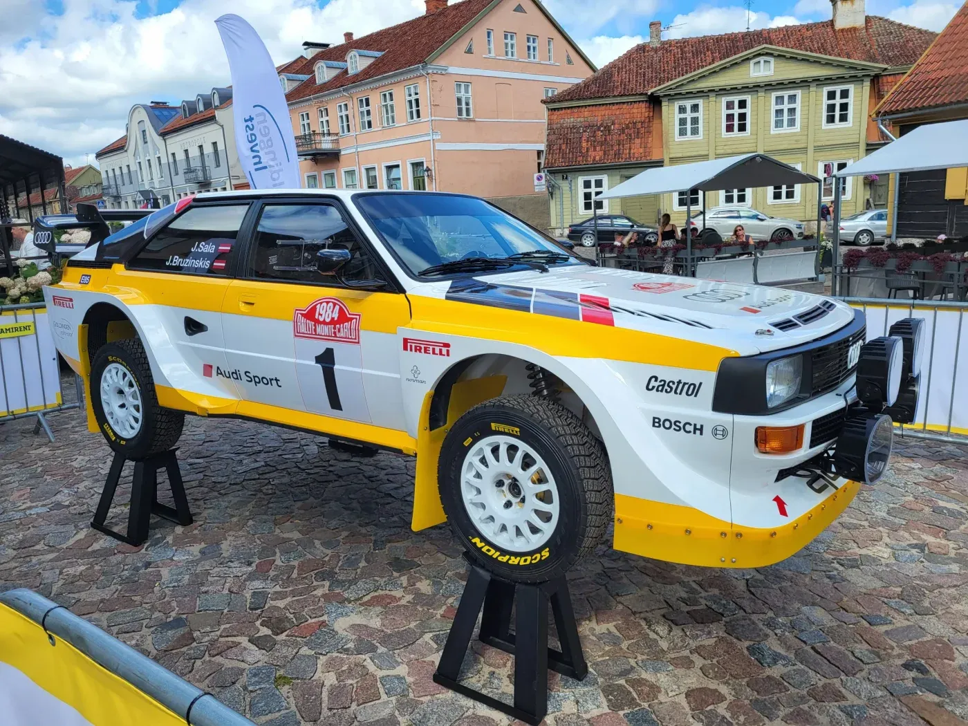 Original photo of a yellow and white Audi rally car on a cobblestone square with buildings in the background
