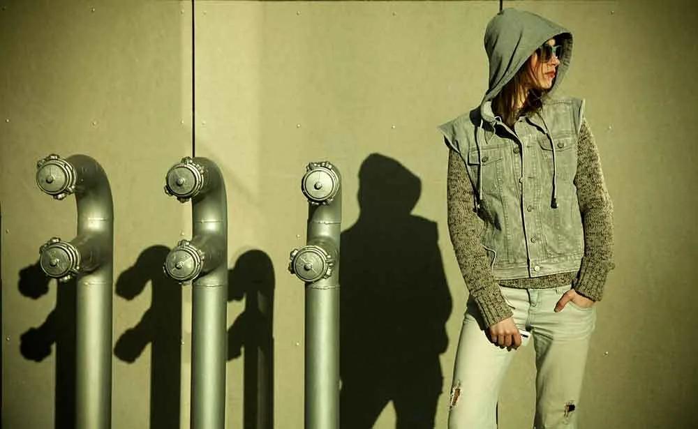 Model in a denim hooded vest posing beside industrial pipes