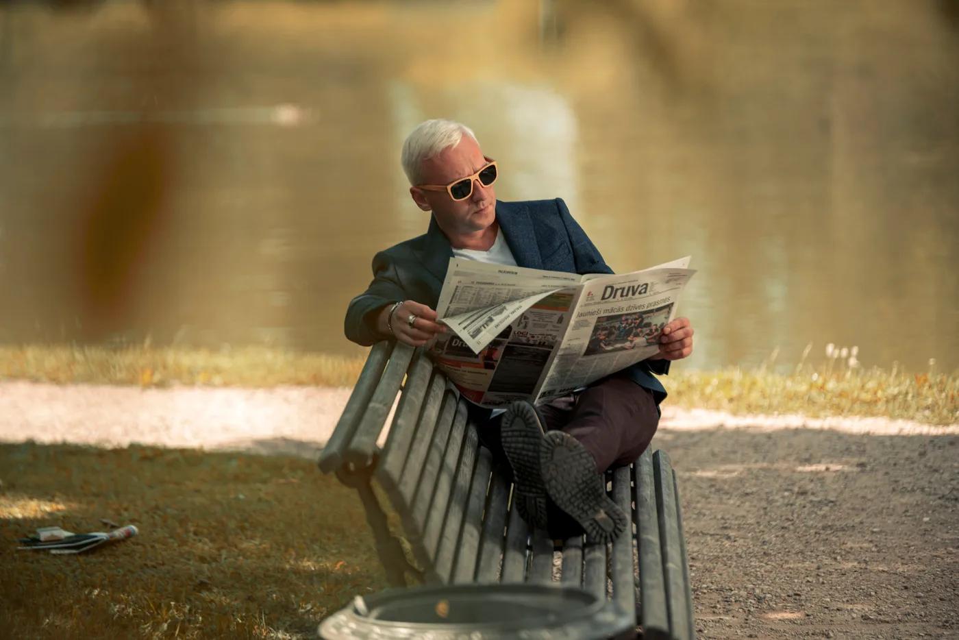 Portrait of a man with white hair and sunglasses reading a newspaper on a park bench
