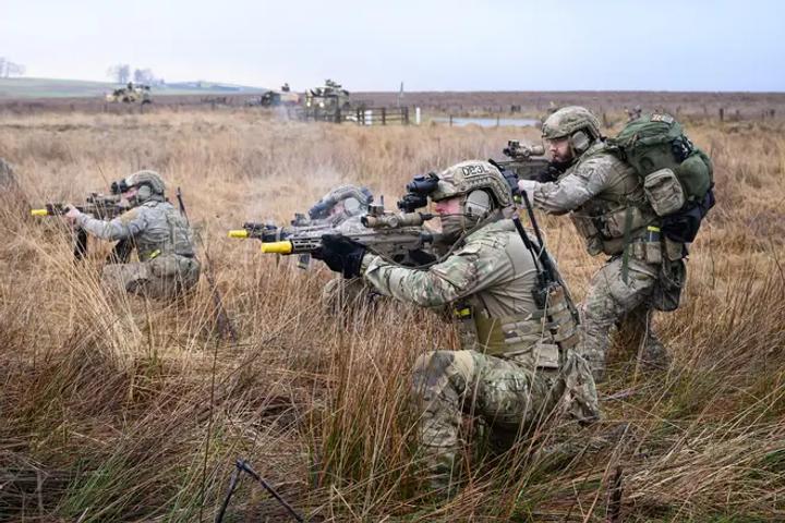 Four soldiers crouch in grass whilst pointing weapons at a target 