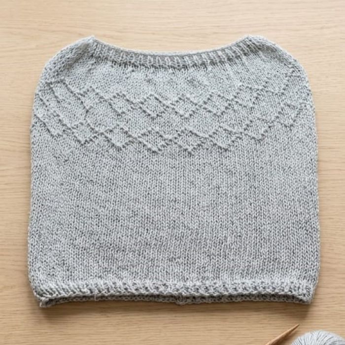 Primer plano de un suéter de lana gris jaspeado en proceso de tejido, con una detallada guarda geométrica de diamantes en el pecho y un borde de punto elástico, exhibido sobre una superficie de madera clara.