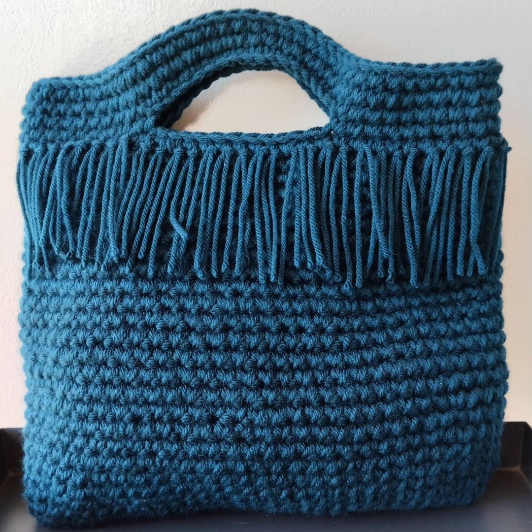 Bolso de mano tejido a crochet en color azul petróleo con detalle de flecos en la parte superior, exhibido sobre una superficie oscura con un fondo minimalista.