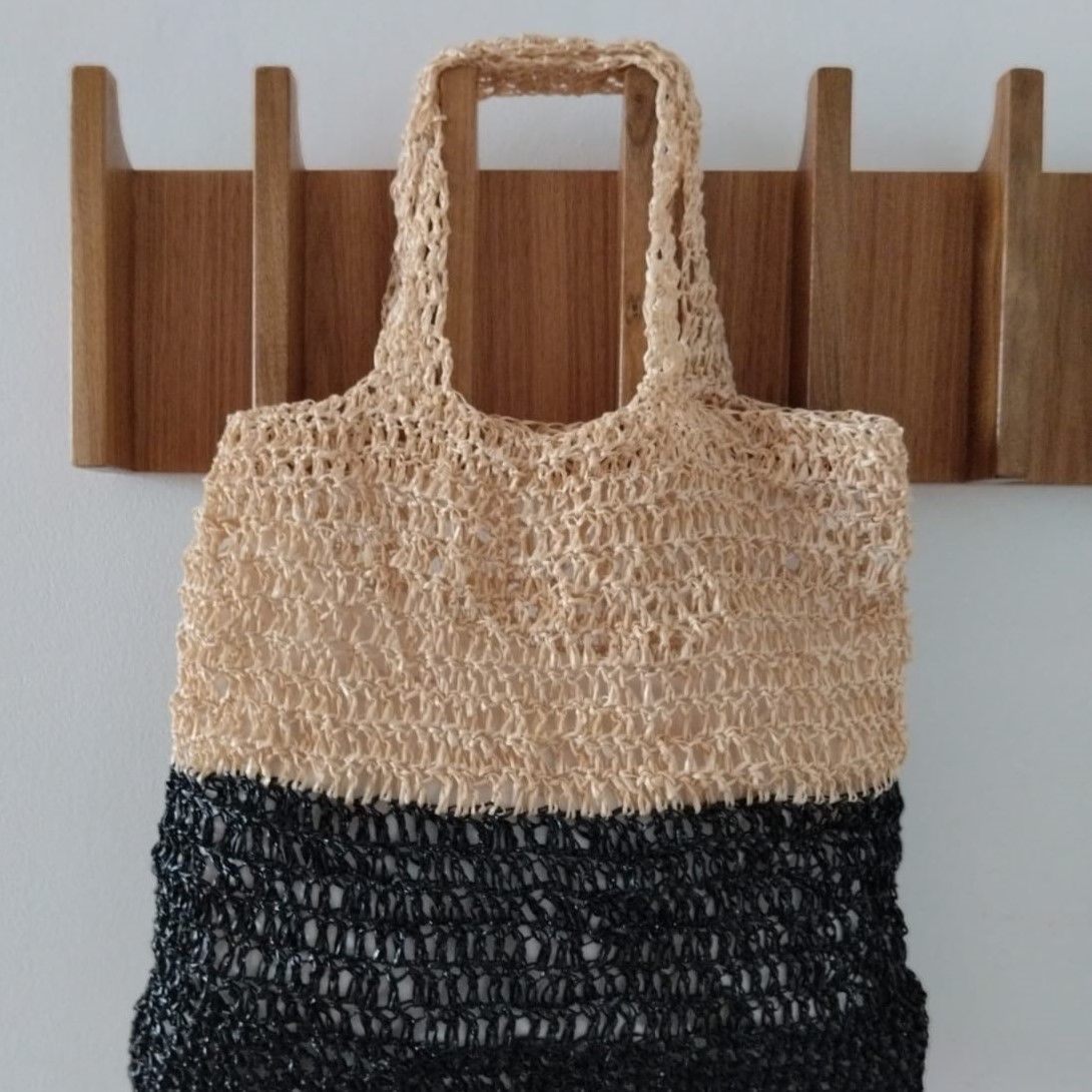 "Bolso artesanal de la colección Arena con diseño bicolor en beige y negro, colgado de un perchero de madera moderno, resaltando la trama calada y rústica del tejido de Nido Trama.