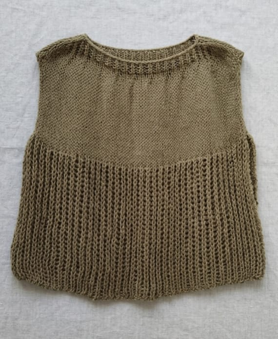 Chaleco de lana tejido a mano en color verde musgo o tierra, con un diseño que combina un panel superior liso y una estructura inferior de punto calado vertical, extendido sobre una base de lino rústico.