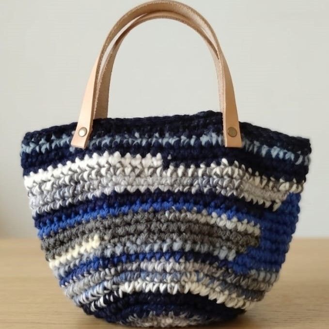 Bolso de mano de ganchillo artesanal con rayas horizontales en tonos azul marino, gris y blanco, con asas de cuero natural y remaches de metal, sobre una mesa de madera.