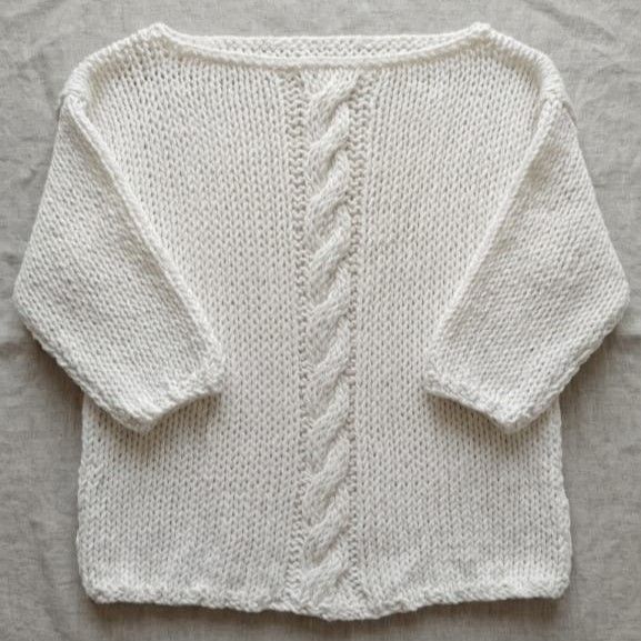 Suéter de lana blanco crudo con mangas tres cuartos y escote barco, destacando un gran diseño de trenza central tejida a mano, sobre un fondo de lino natural.