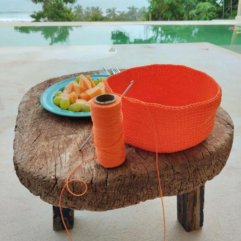 Cesta de crochet color naranja vibrante en proceso de creación sobre una mesa de madera rústica, junto a un ovillo de hilo y un plato de frutas, con una piscina de fondo.