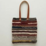 Bolso tote tejido artesanalmente con rayas horizontales en una paleta de colores tierra, bordó y gris, con correas de cuero color suela aseguradas por remaches metálicos.
