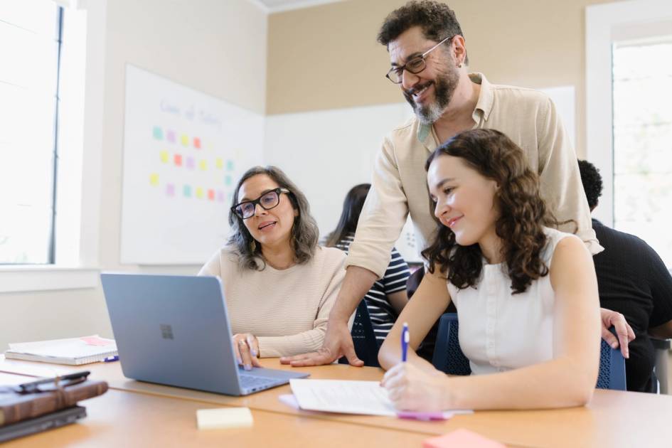 A faculty member leans over to help two graduate students working at a laptop in a bright Rollins classroom, one taking notes as they review material together.