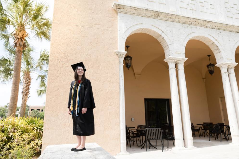 Female college student poses in a cap and gown outside the Bush Science Center.