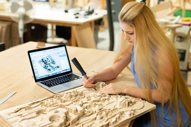 A student cleaning up her topographical CNC wood cutting with a small chisel.