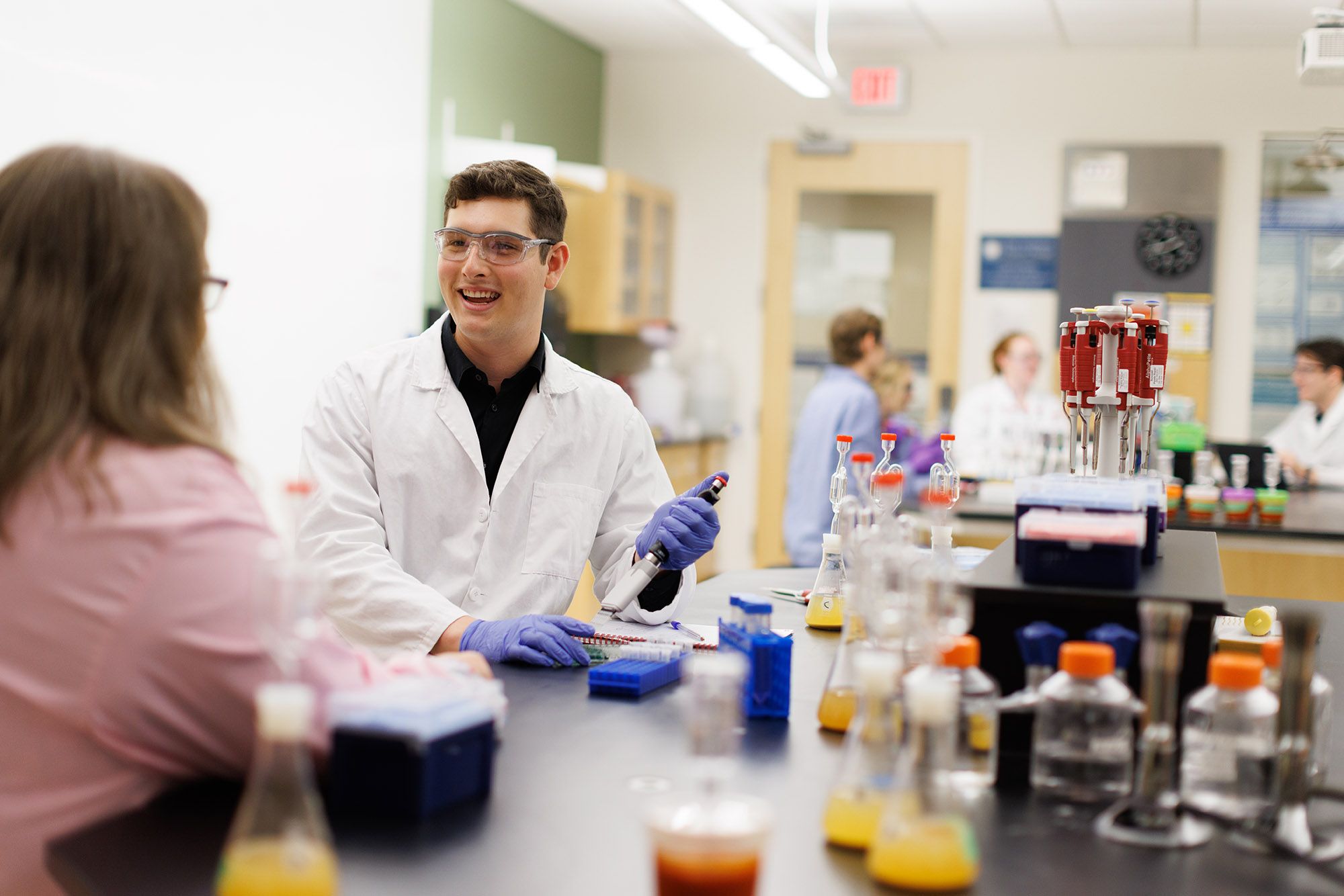 Student in lab coat working with pipette in busy science laboratory.