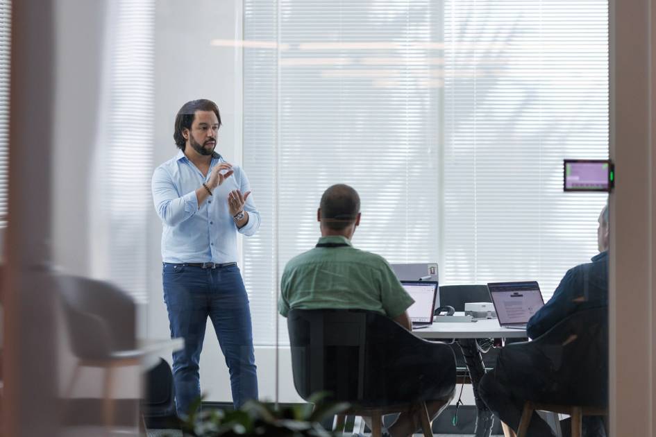 Team meeting in glass-walled office with presenter and laptops visible.
