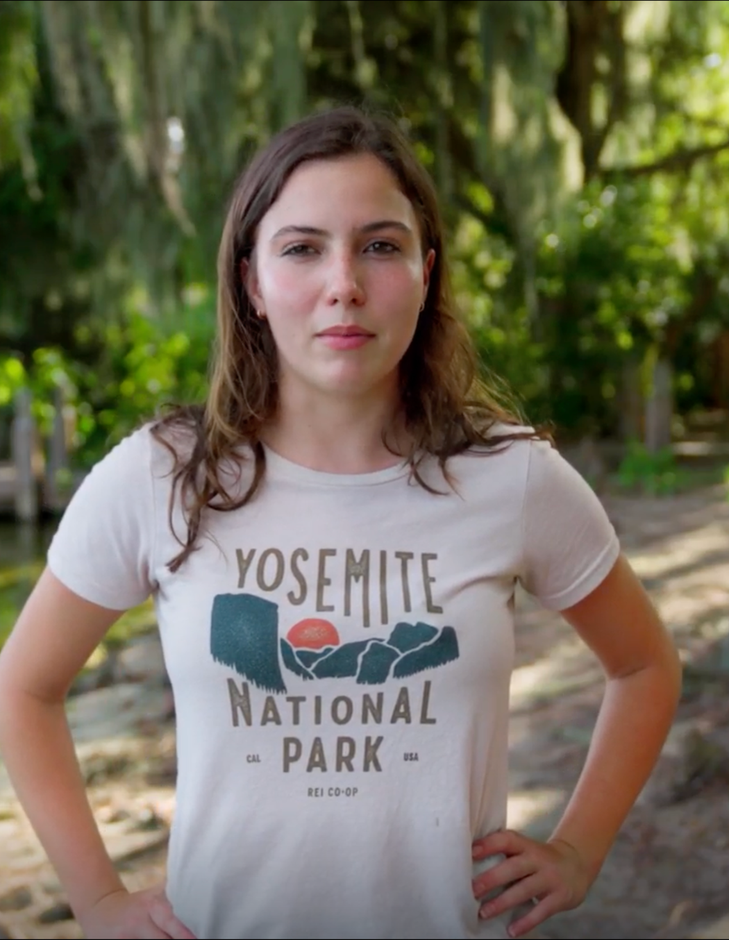 Madi in a white t-shirt that says "Yosemite National Park" standing with hands on hips along the shores of lake with cypress trees.