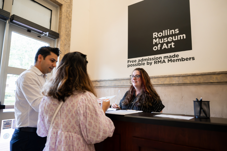 Three people stand indoors, smiling and talking to each other over a desk. One wears a black shirt, smiling at the others who are reading a piece of paper. In the background, a large logo is painted on the wall that reads "Rollins Museum of Art."