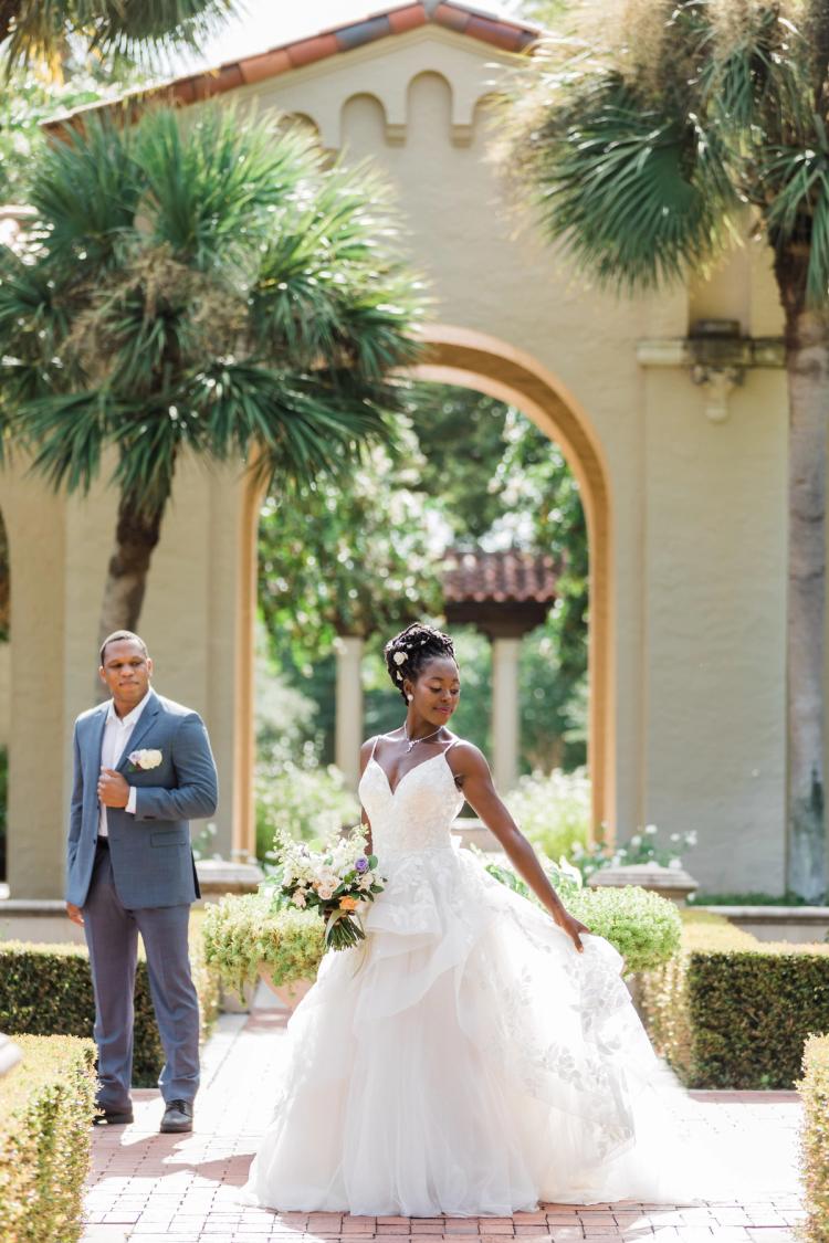 A bride and groom posing in a garden in front of a stone archway framed by palm trees