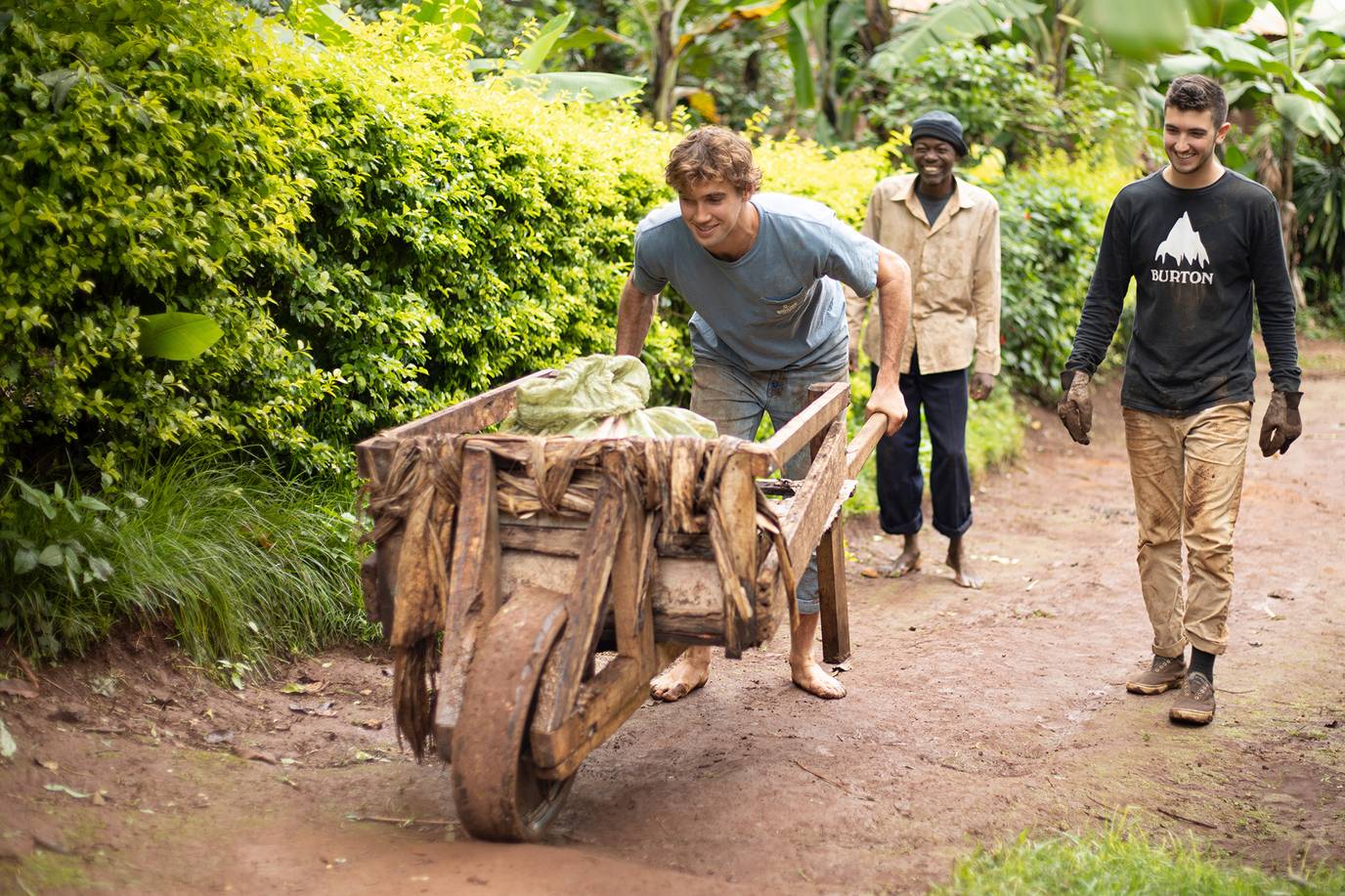 A college student pushing a wooden wheelbarrow barefoot along a dirt path.
