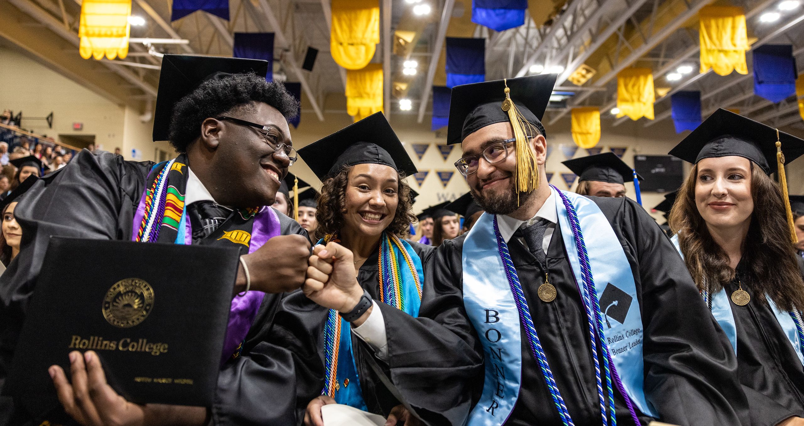 Students in graduation robes, fist-bumping and smiling.