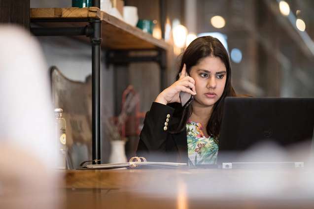 A college alumn sitting in a cafe on her computer while listening to her phone.