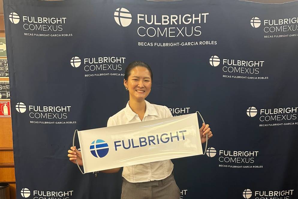 A student stands in front of a Fulbright banner and with a Fulbright sign