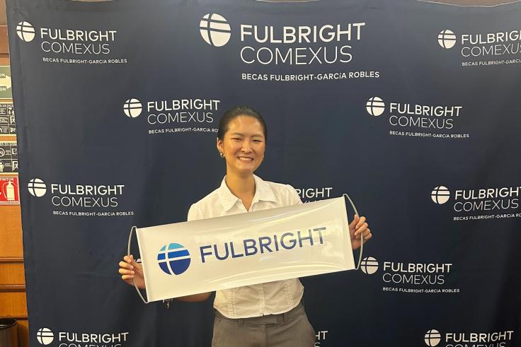 A student stands in front of a Fulbright banner and with a Fulbright sign