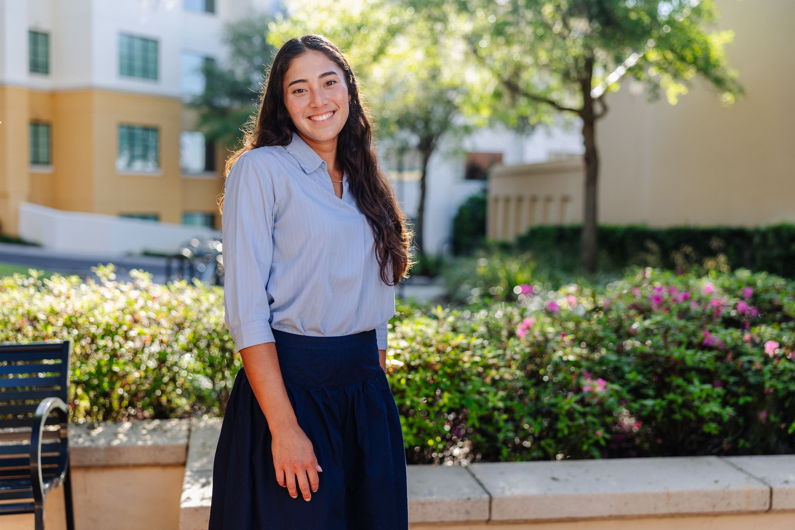 Smiling student in a light blue blouse and navy skirt stands outdoors beside a flower bed, with campus buildings behind her.