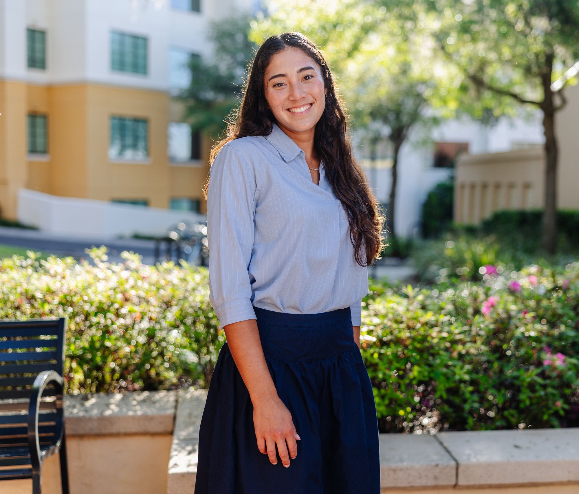 Smiling student in a light blue blouse and navy skirt stands outdoors beside a flower bed, with campus buildings behind her.
